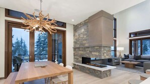A modern living room with a stone fireplace, a dining area, and large windows offering a view of trees. - Lakeside townhouse w/ private garage (Whistler)