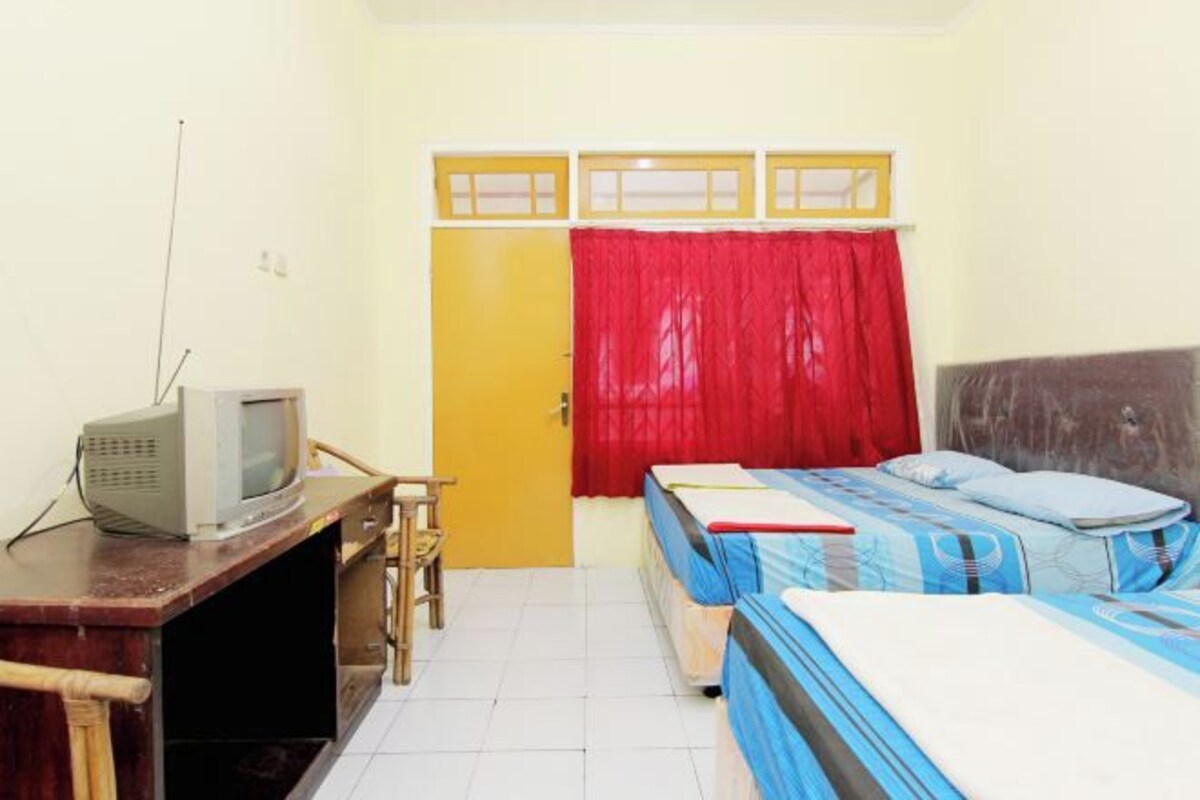 Una habitación con dos camas, un escritorio con televisor, una puerta y una ventana con cortina roja.