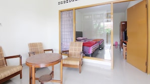 Una habitación con una mesa redonda de madera, dos sillas de mimbre, una cama con ropa de cama colorida y un ventanal corredero que da a otra habitación.