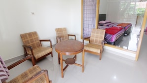 Una habitación con una mesa redonda de madera rodeada por cuatro sillas de mimbre, un espejo grande que refleja una cama con colcha a rayas y una ventana con cortinas traslúcidas.