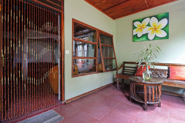 A room with a wooden ceiling, a large window with multiple panes, a wooden bench, a round wooden table with a vase, and a wall-mounted painting of flowers. - Bagus Bay Guest House (Samosir)
