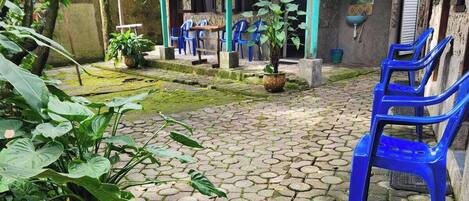 A courtyard with hexagonal stone paving, a covered seating area with blue plastic chairs, and a potted plant.