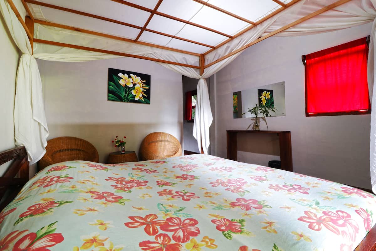 A bedroom with a floral bedspread, a wooden headboard, a bedside table with a vase, and a red window blind.