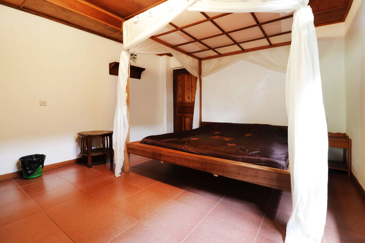 A single bed with a wooden frame and a canopy, a small wooden table, a green trash bin, and a door in a room with tiled flooring.