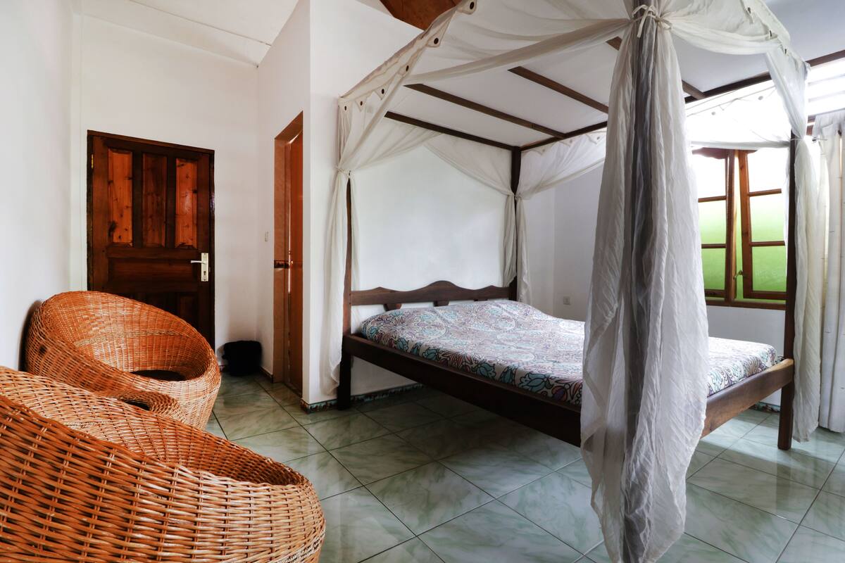 A room with a bed under a canopy, a wicker chair, and a wooden door.