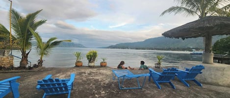 A waterfront seating area with blue chairs and potted plants, overlooking a body of water and distant mountains.