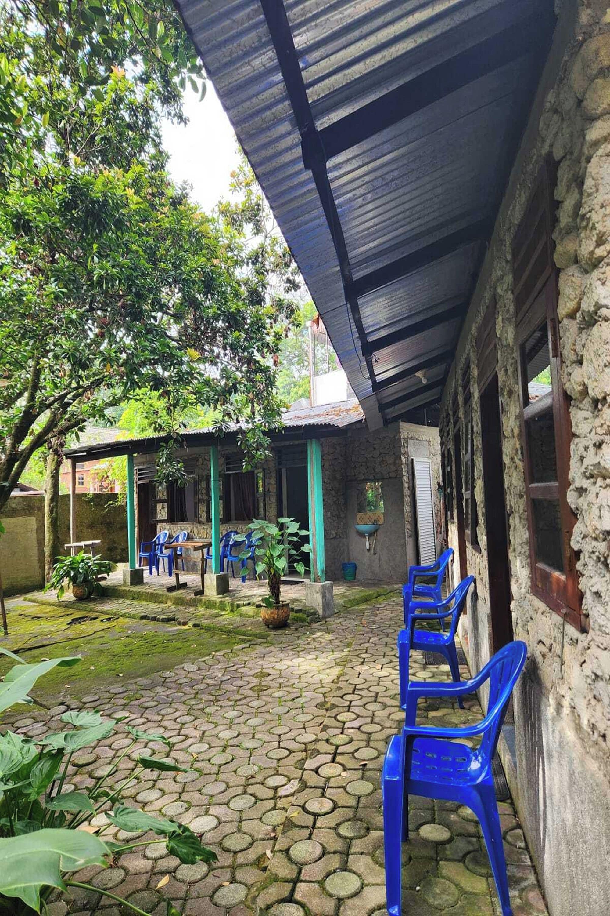 Ein Innenhof mit einem pflasterbelagten Bereich, einer überdachten Sitzgelegenheit mit blauen Plastikstühlen und einem Gebäude mit einer wellenförmigen Metalldach.