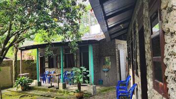 Un patio con área empedrada de piedra, zona de estar techada con sillas de plástico azul y un edificio con techo de lámina corrugada.