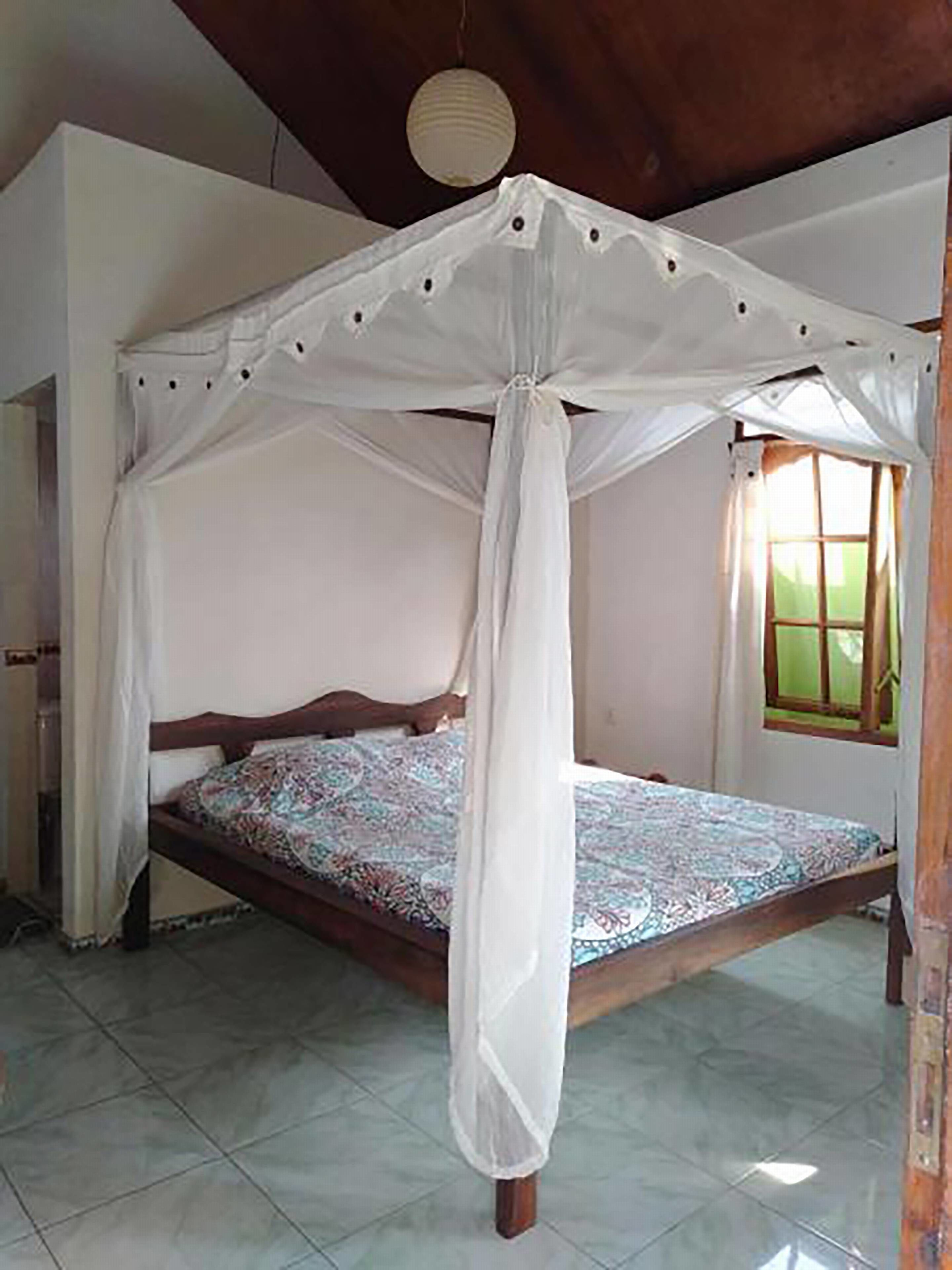 Une chambre avec un lit sous un baldaquin blanc, une fenêtre avec des rideaux et un sol carrelé.