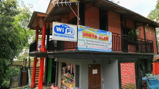 A two-story building with a red brick facade, a balcony with a red railing, and signs advertising free WiFi and a karaoke bar. - Penginapan Villa Surya Alam (Prigen)