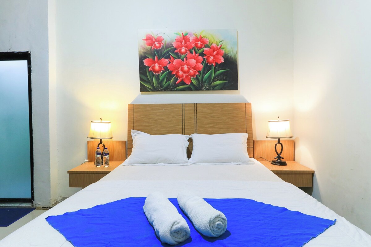A hotel room with a bed, two bedside lamps, a painting of flowers, and a blue blanket.
