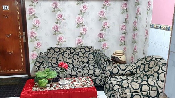A living room with patterned sofas, a red table with a vase and plant, and a door marked with the number 08. - Dharmawati Homestay (Sidoarjo)