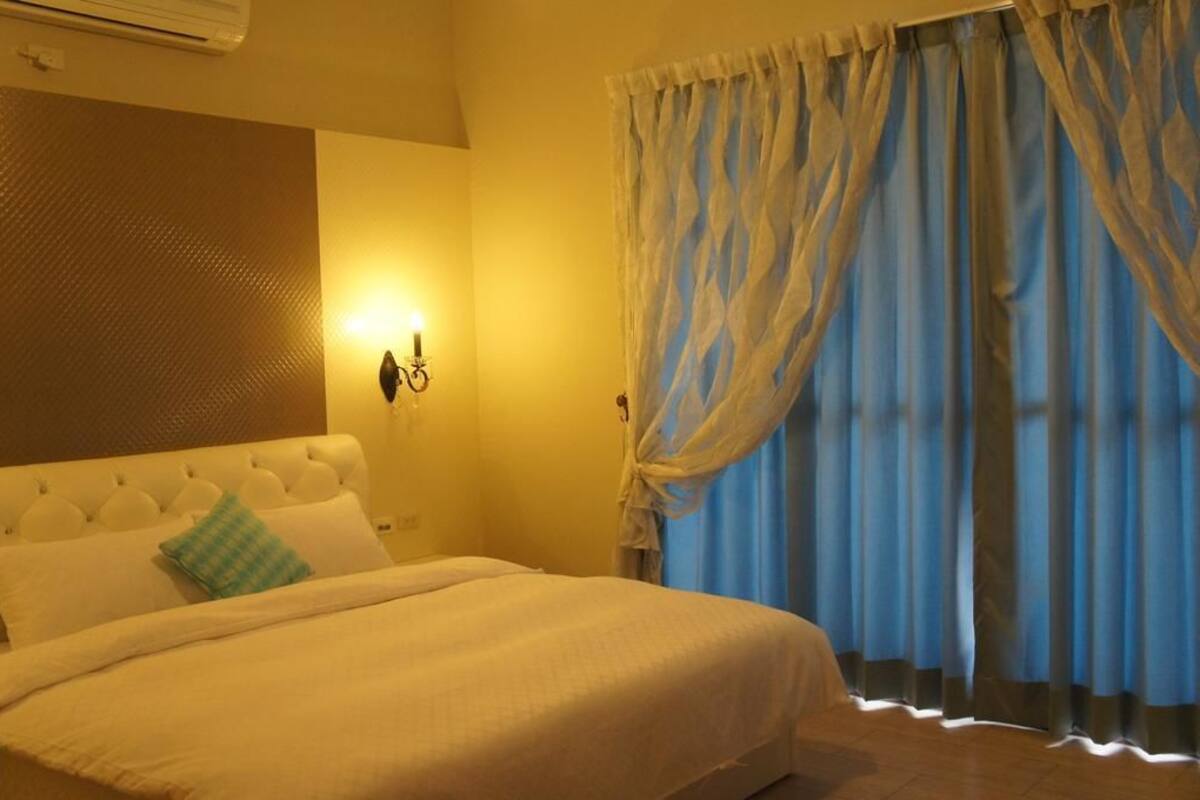 A hotel room with a large bed, a wall-mounted air conditioner, a wall sconce, and a window with blue curtains.