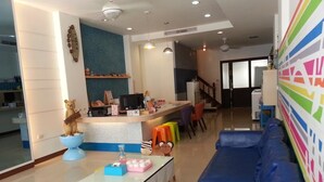 A modern living room with a blue sofa, a glass coffee table, and colorful stools. There is a kitchen area with a refrigerator and a microwave. - Just Home (Magong)