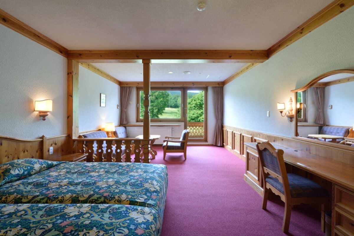 Une chambre d’hôtel avec un lit, un bureau, une chaise et une vue sur un jardin, visible par une grande fenêtre.