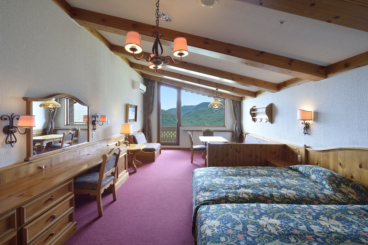 Une chambre avec un lit en bois, un bureau avec une chaise, une commode et une grande fenêtre donnant sur les montagnes.