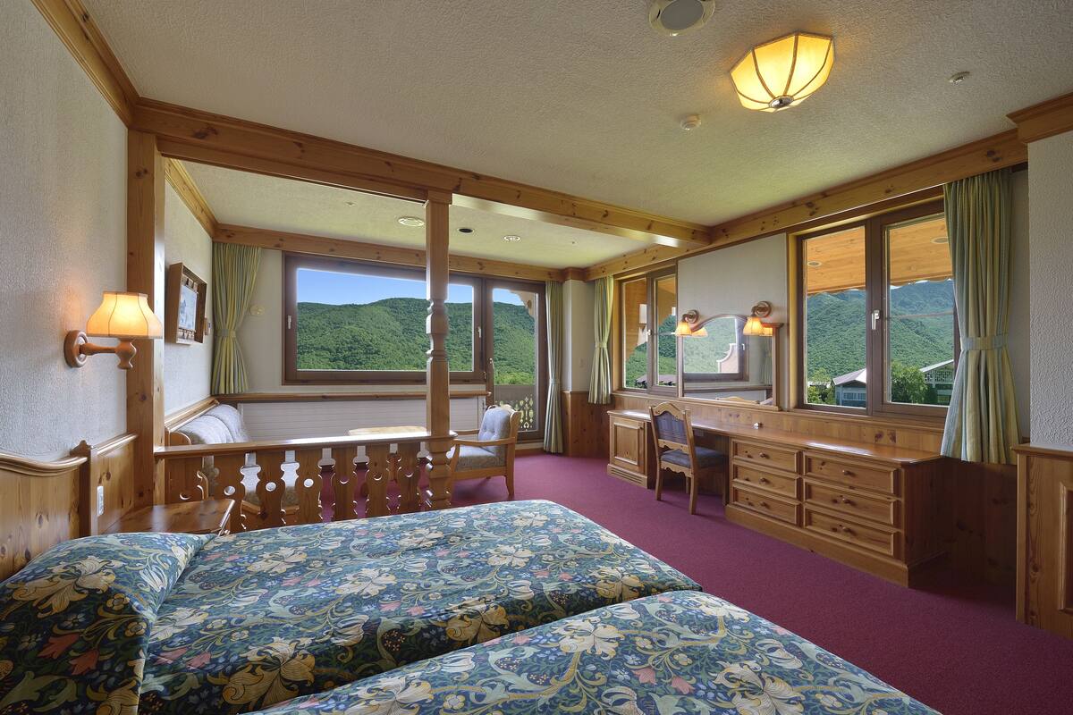 Une chambre avec deux lits, des meubles en bois, un bureau et de grandes fenêtres donnant sur des collines verdoyantes.