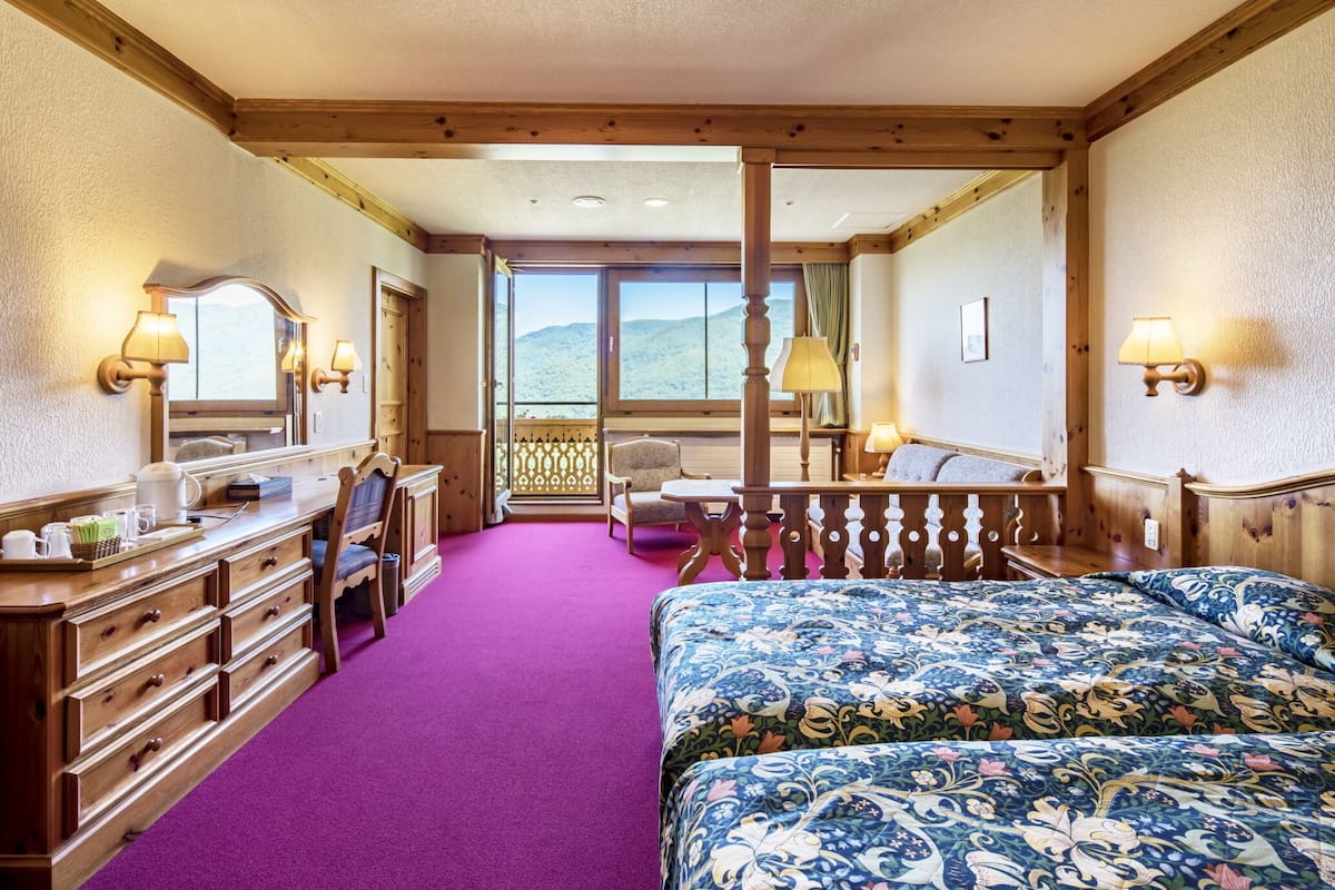 Une chambre d’hôtel avec un lit en bois, une commode, un miroir et une vue sur les montagnes, visible à travers de grandes fenêtres.