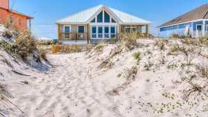 Une maison située en bord de mer, dotée d’une grande véranda, de nombreuses fenêtres et d’une plage de sable au premier plan.