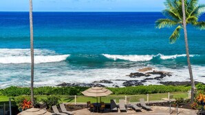 Une vue sur la plage, avec les vagues de l’océan, des palmiers et une terrasse dotée de chaises longues et de parasols.