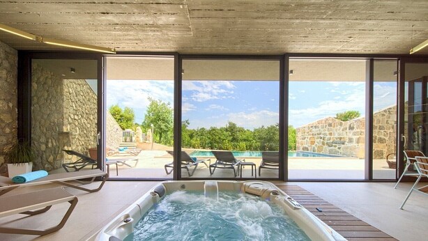 A modern indoor swimming pool area with a hot tub, lounge chairs, and a view of an outdoor pool and greenery.
