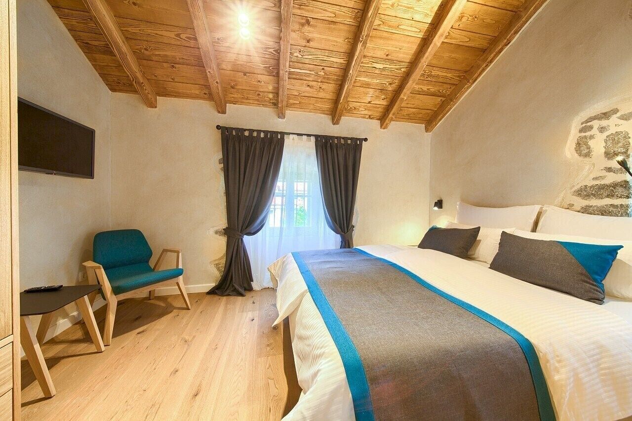 A bedroom with a wooden ceiling, a bed with a blue and white bedspread, a chair, and a window with curtains.