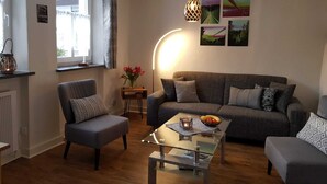 Un salon chaleureux avec un canapé gris, deux fauteuils, une table basse en verre et une petite table d’appoint sur laquelle est posé un vase de fleurs.