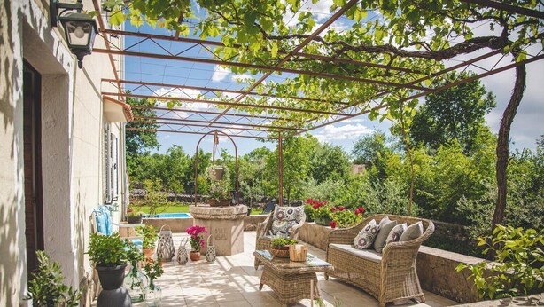 Une terrasse couverte, aménagée avec des meubles en osier, des plantes en pot et une fontaine en pierre.