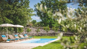 Un espace piscine aménagé avec des chaises longues et des parasols, entouré de verdure et délimité par un mur de pierre.