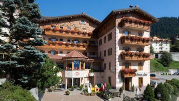 Un hotel con numerosi balconi decorati con fiori, un ingresso centrale con tetto ornamentale e un bar snack nelle vicinanze.