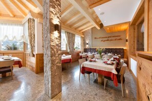 A dining area with multiple tables set for meals, stone walls, wooden beams, and a view of the outdoors. - Hotel Cristallo (Andalo)