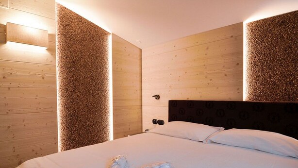 A hotel room with a wooden wall, a bed with white linens, and a headboard with a pattern. - Hotel Cristallo (Andalo)