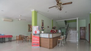 Ein Hotel-Empfangsbereich mit einem hölzernen Empfangstresen, einem roten Tisch mit Stühlen, einem Deckenventilator und einer Treppe im Hintergrund.