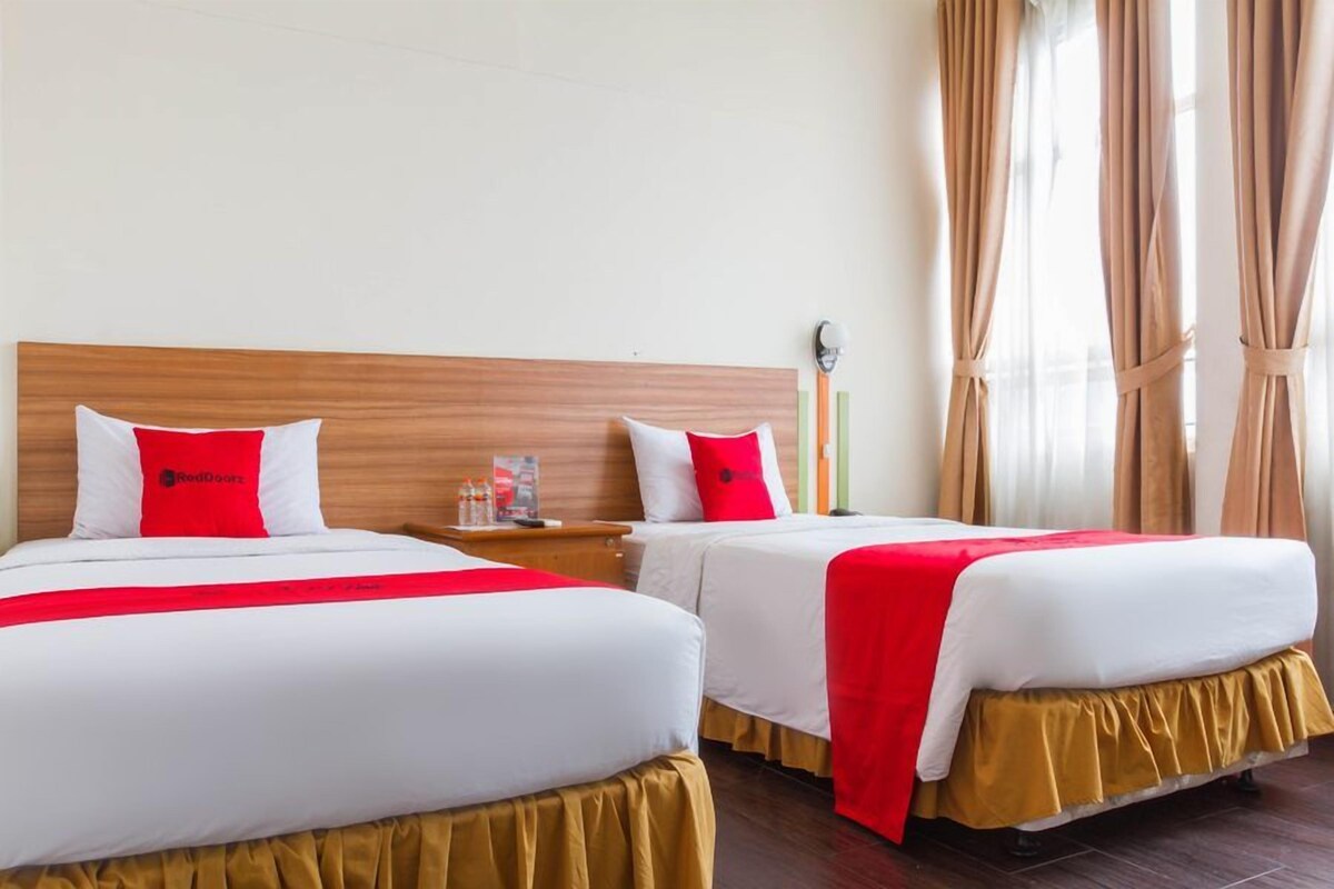 A hotel room with two beds, each with white bedding and red accents, a wooden headboard, a nightstand with a lamp, and a window with curtains.