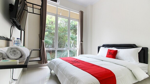 A hotel room with a bed, a TV, a window with curtains, and a view of greenery outside. - RedDoorz near Jogja City Mall 3 (Pogung Lor)