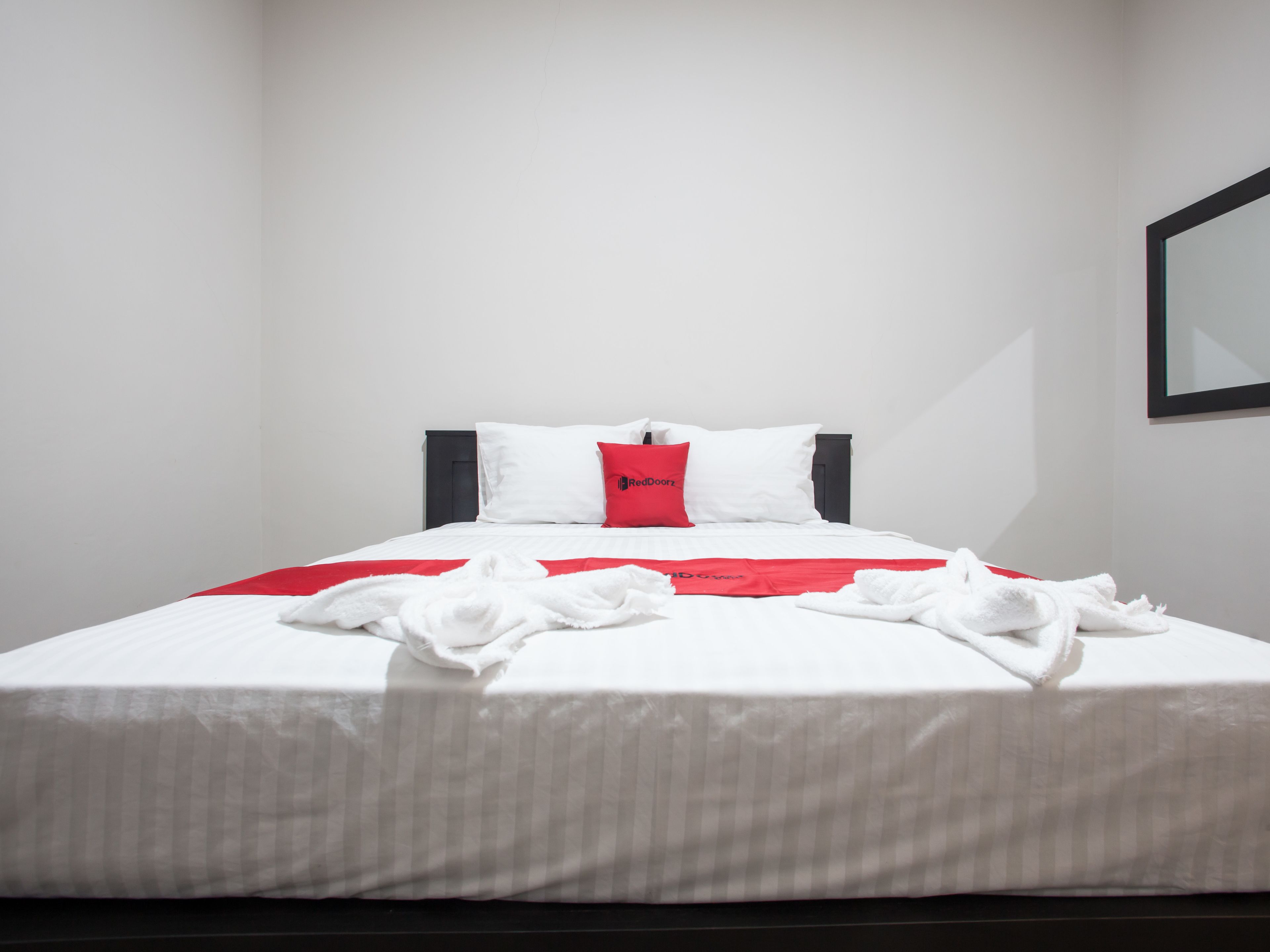 Un lit bien fait, avec une literie blanche et un coussin rouge qui apporte une touche de couleur, une tête de lit noire et un miroir encadré accroché au mur.