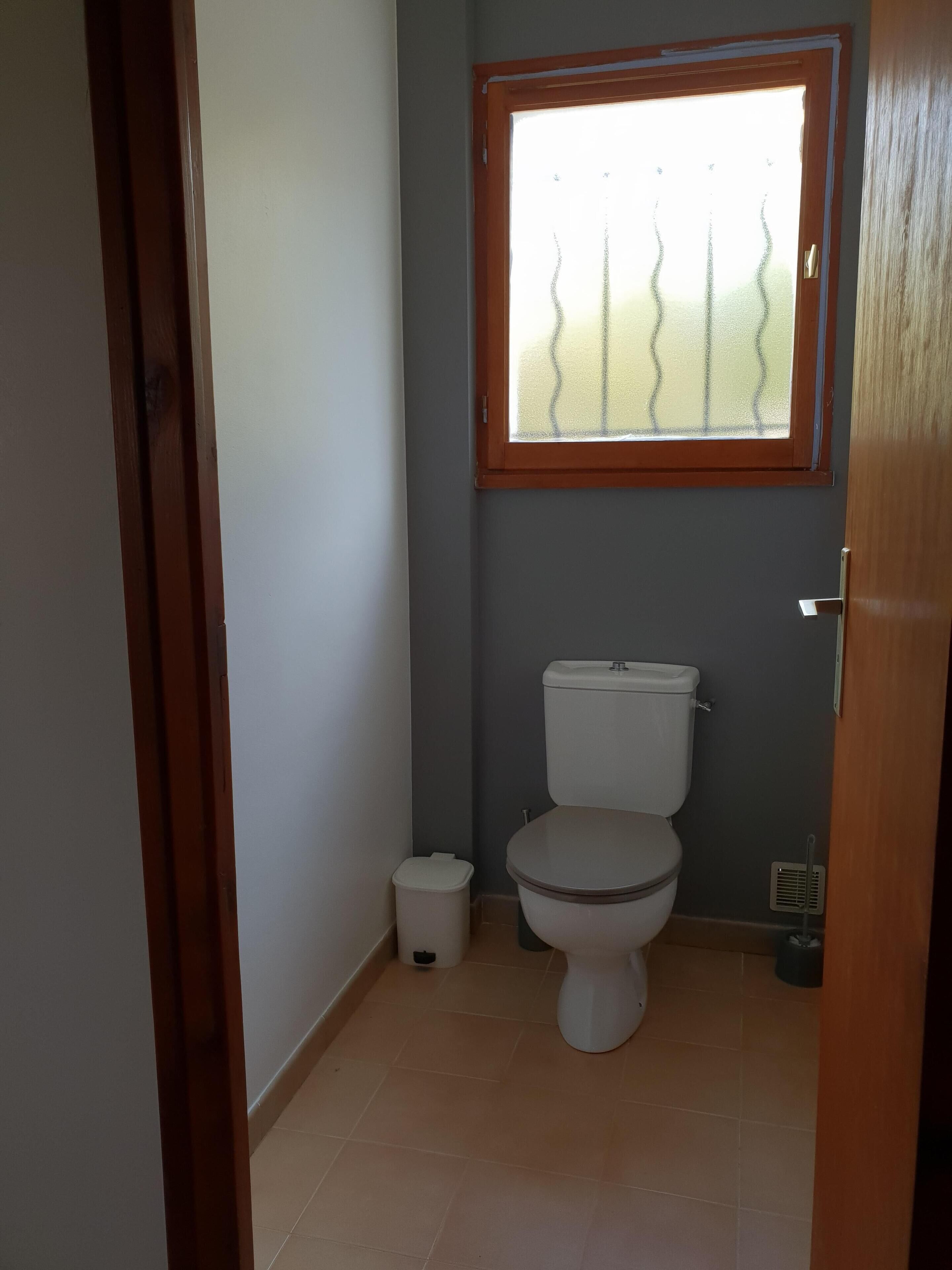 Une petite salle de bain comprenant des toilettes, une poubelle et une fenêtre avec un cadre en bois.