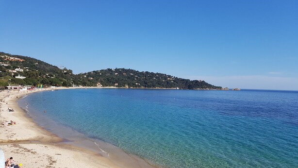 A beach with clear blue water, sandy shore, and buildings on a hillside in the background.