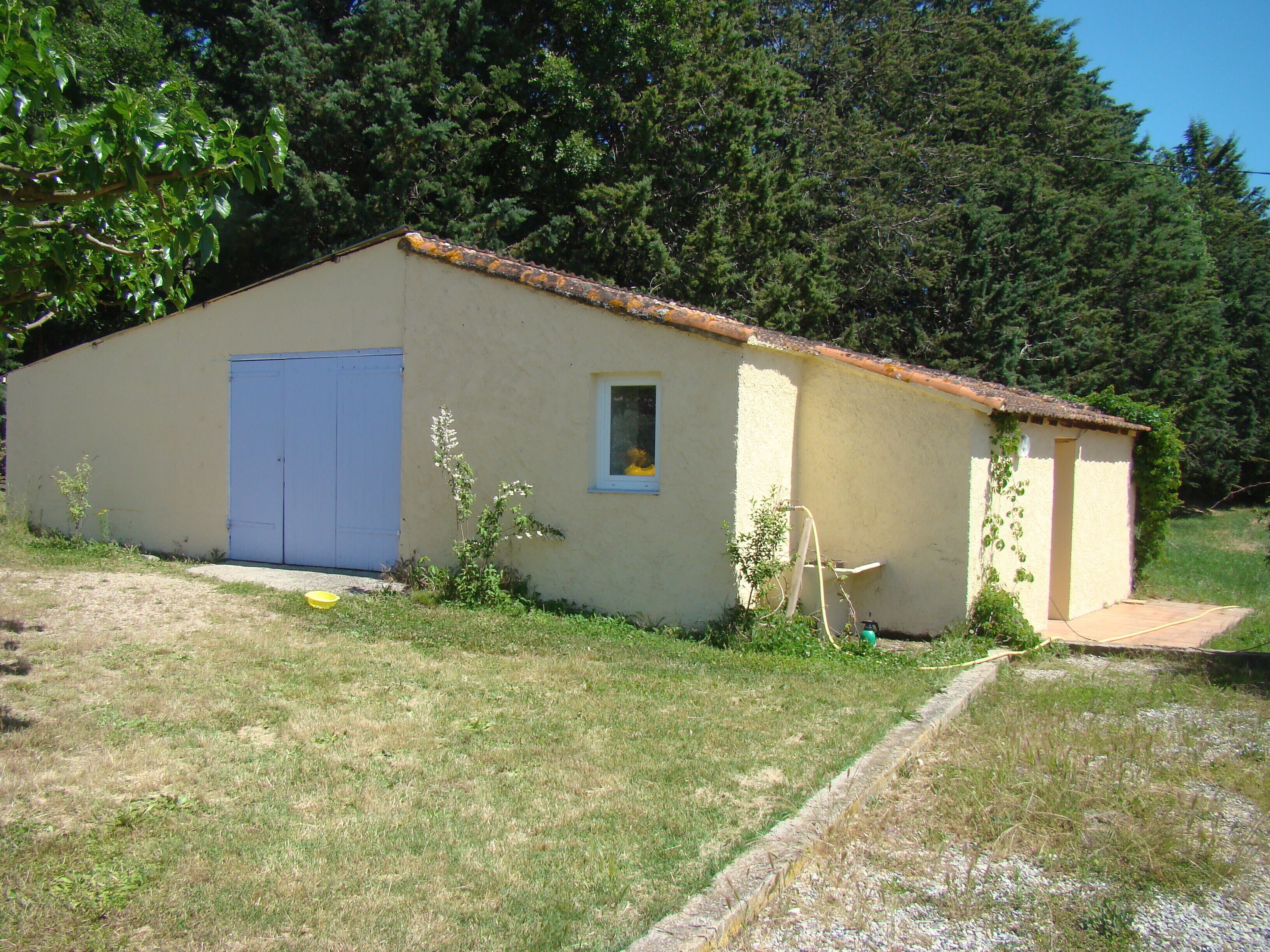 Un piccolo edificio a un piano con una porta blu e una piccola finestra, immerso nel verde tra l'erba e gli alberi.