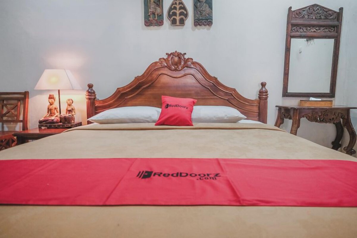 A bedroom with a wooden headboard, a bed with a red pillow, a bedside table with a lamp, and a mirror on the wall.