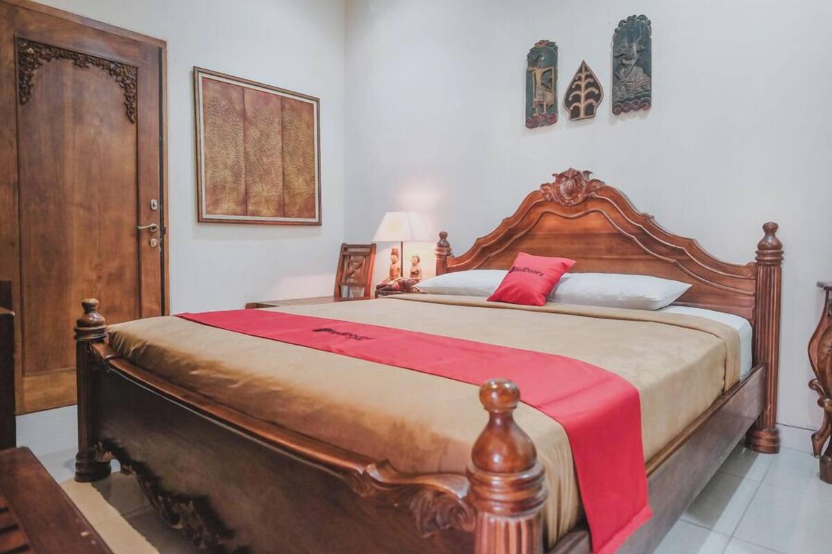 A wooden bed with a red blanket, a wooden door, a framed artwork, and a wall-mounted decorative piece.