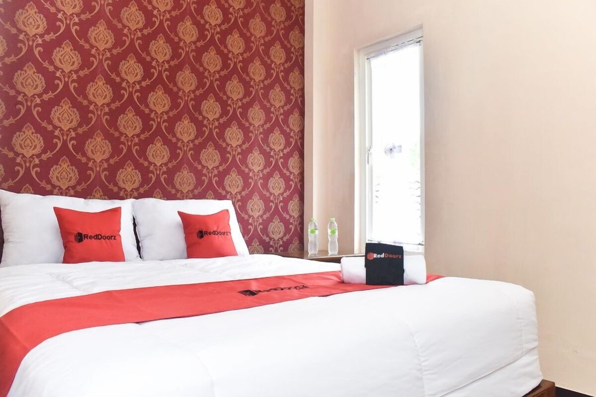 A hotel room with a patterned wallpaper, a bed with white bedding and red accents, a window, and a RedDoorz branded item on the bed.