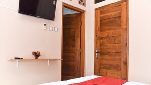 A hotel room with a bed, a television mounted on the wall, a wooden door, and a small shelf with a plant. - RedDoorz Syariah near Batu Night Spectacular (Batu)