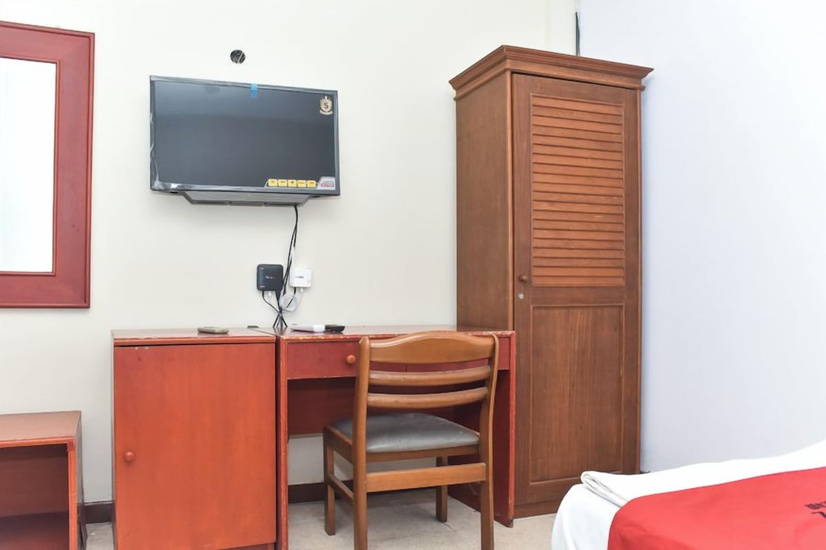 A room with a wooden desk, a television mounted on the wall, a wooden wardrobe, and a bed with a red blanket.