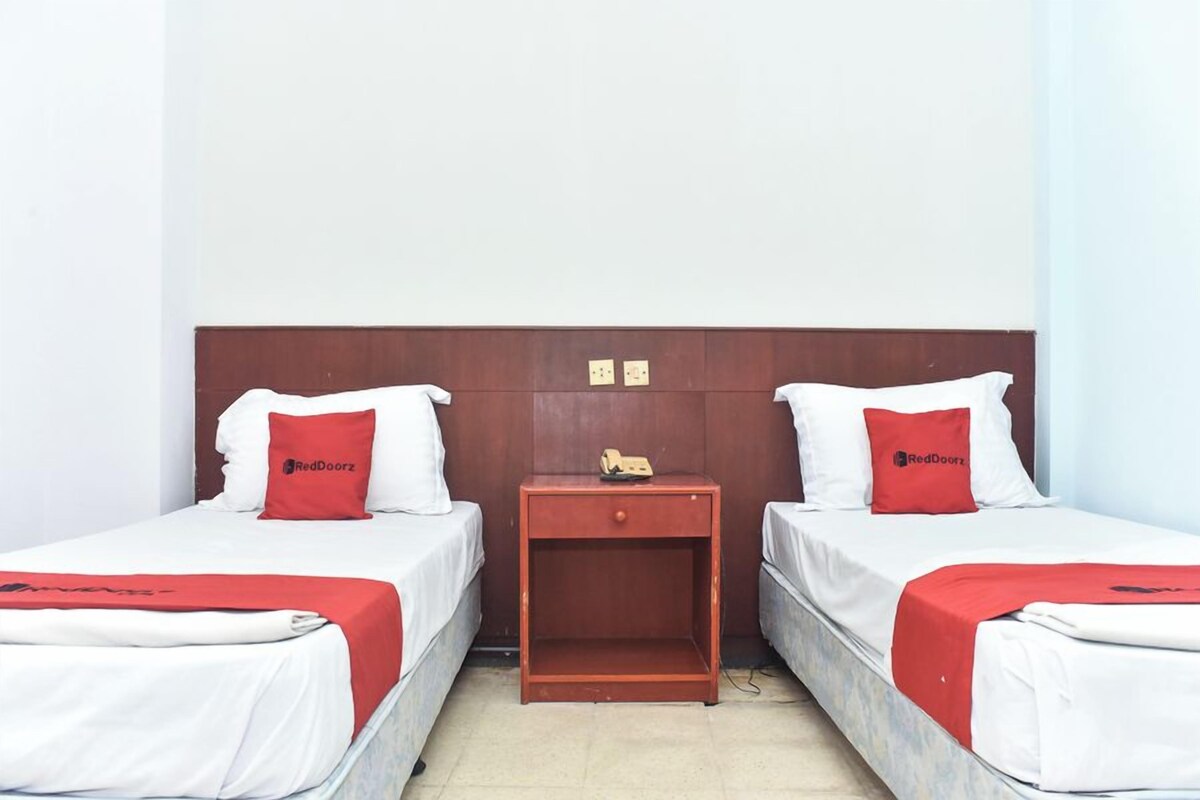 Una habitación de hotel con dos camas individuales, una mesita de noche de madera y una colcha roja y blanca.