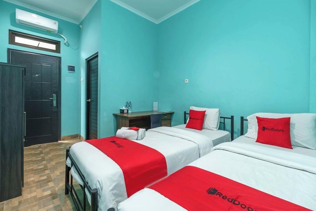 A hotel room with two beds, each with a red and white bedspread and a pillow with the RedDoorz logo. There is a desk with a chair, a small table with a bottle, and a wall-mounted air conditioner.