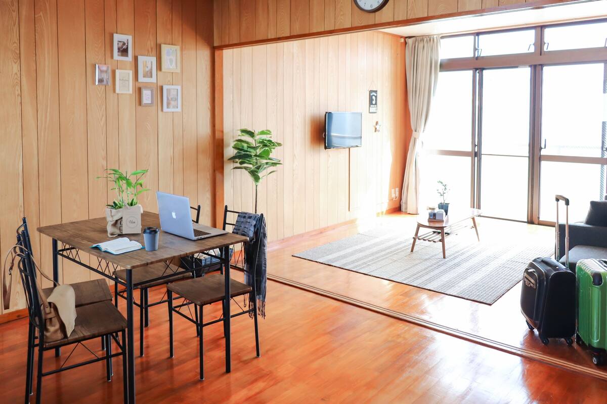 A wooden-paneled room with a dining table, chairs, a laptop, a plant, a TV, and luggage.