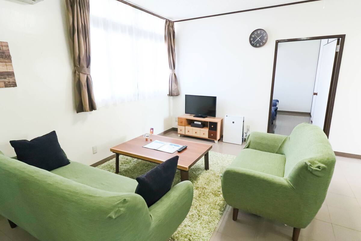 A small living room with two green sofas, a wooden coffee table, a flat-screen TV on a wooden stand, and a small cabinet.