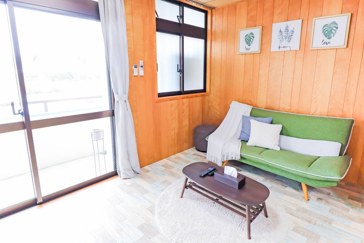 A room with a green sofa, a remote control on a coffee table, and a window with curtains.