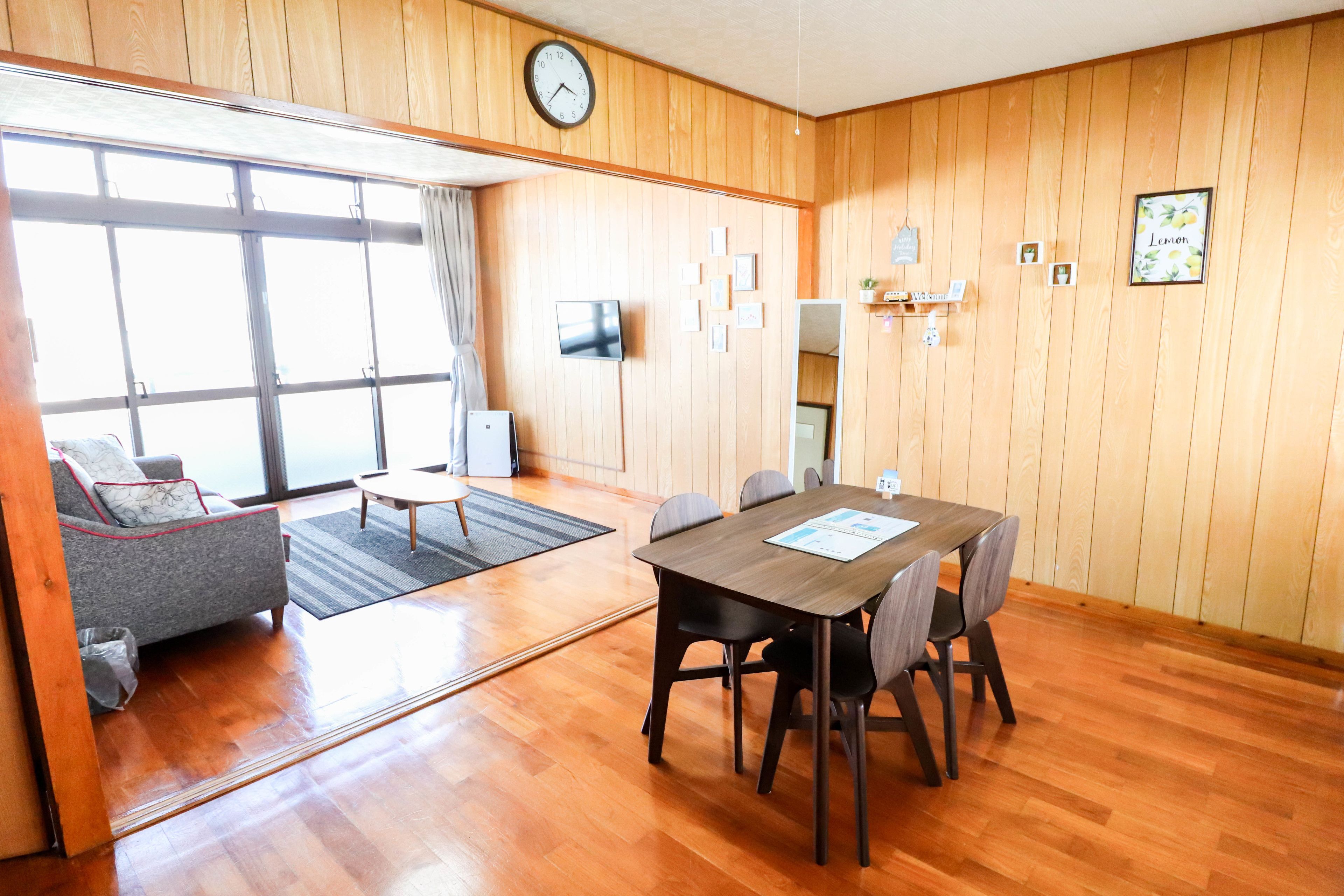 Una habitación con piso de madera, una mesa de comedor con sillas, un sofá, un televisor y un reloj en la pared.